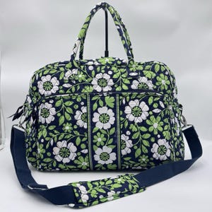May include: A quilted duffel bag featuring a navy blue background with a repeating pattern of white and light green flowers and leaves. The bag includes two top handles, a detachable navy blue shoulder strap, and a front zippered pocket.