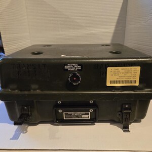 May include: A dark green rectangular transit case with a hinged lid and metal latches. The case has several labels and text, including "TRANSIT" and "RELIEF VALVE." A small valve is visible on the front. The case appears to be military or industrial equipment.