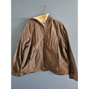 May include: Brown hooded jacket with a yellow interior lining, a zipper closure, and two front pockets. The jacket is hanging on a metal hanger against a blue-gray wall. The jacket appears to be made of a durable, possibly water-resistant material.