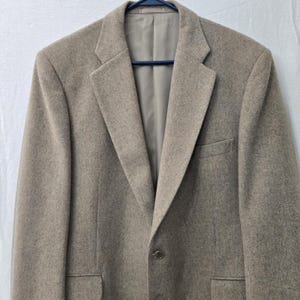 May include: A tan blazer with a two-button closure, notched lapels, and a chest pocket. The jacket is hanging on a blue hanger against a white background. The blazer is made of a textured fabric.