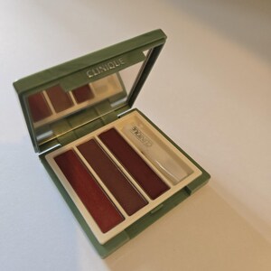 May include: Open olive green Clinique eyeshadow palette with a mirror. The palette contains four shades of red and brown eyeshadow. The brand name "Clinique" is printed on the palette.