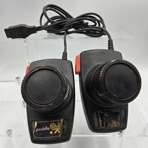 May include: Two vintage black paddle controllers with red buttons and large dials. The controllers have a black cable and connector. One controller has a sticker that says "paddle" with a tennis ball and racket graphic.