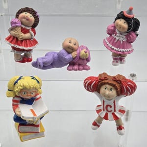 May include: A collection of Cabbage Patch Kids figurines. One holds an ice cream cone, another is dressed for a party, and a third is reading a book. The other two figurines are a baby with a dog and a cheerleader.
