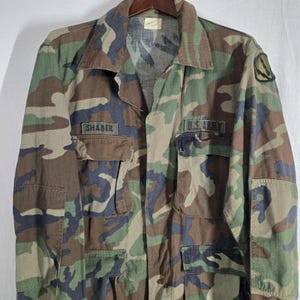 May include: Camouflage pattern military jacket with the text "SHABER" above the left pocket and "U.S. ARMY" on the right. The jacket features a button-down front, two chest pockets, and a patch on the right sleeve. The colors include green, brown, and black.