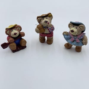 May include: Three decorative bear figurines. One bear is seated with a paddle, another holds a honey jar, and the third wears a dress. The bears are painted in shades of brown, red, blue, and yellow, with detailed facial features and clothing.