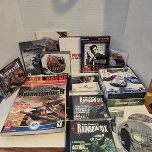 May include: A collection of PC game boxes and CDs, including titles like "Max Payne," "Tom Clancy's Rainbow Six," and "Medal of Honor: Breakthrough." The games feature action-oriented themes and military strategy elements, with various box art designs.