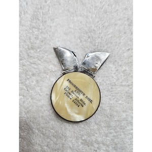 May include: Vintage silver-toned metal pin with a circular, off-white center. The pin features the text "PROGRESSIVE DIST. LTD." and address details. The pin has two wing-like extensions at the top.