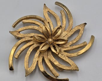 May include: A gold-toned metal brooch in a floral design. The brooch features a central flower with detailed petals, surrounded by curved, petal-like elements. The brooch has a textured surface and a pin closure on the back.