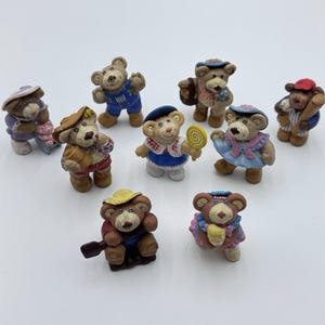 May include: A collection of small, decorative bear figurines. Each bear is uniquely dressed in outfits like overalls, hats, and dresses, some holding accessories such as a lollipop or a baseball bat. The bears are painted in various colors, creating a charming and whimsical display.