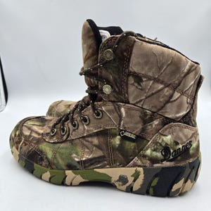 May include: A Danner camouflage print hiking boot featuring a high-top silhouette, brown and green earth-tone patterns, metal eyelets, dark brown laces, a black Gore-Tex tag on the side, and a thick, textured rubber sole with a matching camo design.