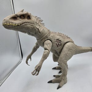 May include: A light gray dinosaur action figure with a textured, scaly skin surface, sharp yellow teeth, and orange eyes. The figure features a prominent row of spikes along its back and a visible three-slash battle damage mark on its left side.