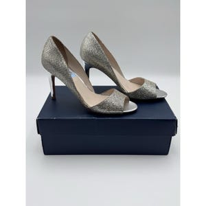 May include: A pair of silver glitter peep-toe heels with silver heels. The shoes are displayed on a dark blue box. The heels have a classic design, suitable for formal events or special occasions.