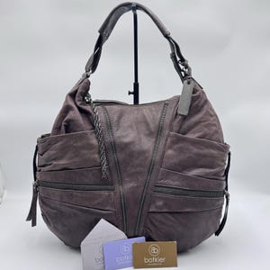 May include: A dark gray hobo bag with a slouchy silhouette, featuring multiple diagonal zippered pockets, a single shoulder strap, and a hanging metal tag. Three small rectangular cards with the Botkier logo are positioned at the base.