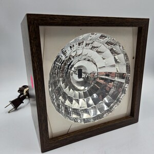 May include: A vintage, square-framed light fixture with a faceted glass bulb. The dark brown wooden frame surrounds a white interior, and a brown electrical cord with a plug extends from the side. The bulb is clear and reflects light.