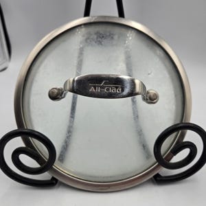 May include: A stainless steel All-Clad pot lid with a clear glass center. The lid has a rounded handle with the brand name engraved on it. The lid is displayed on a black metal stand.