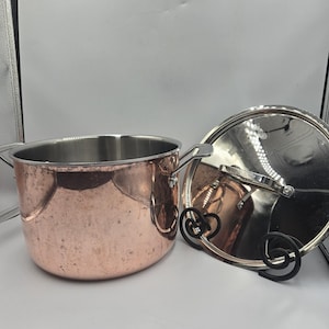 May include: A copper-colored cooking pot with a stainless steel interior and two side handles. The pot is next to a stainless steel lid with a black handle. The pot and lid are on a white surface.
