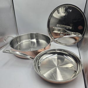 May include: A set of stainless steel cookware with copper-colored exteriors. The set includes a frying pan with a long handle, a saucepan with two small handles, and a lid with a stainless steel handle. The cookware is displayed on a white surface.