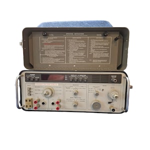 May include: An open, olive-drab metal case with a blue padded top, revealing a Hewlett-Packard transmission test set. The set features numerous dials, switches, and input jacks, with text including "DISPLAY & MONITOR" and "INTERNAL MOD. & OHM TERM. NO."