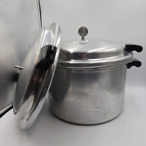 May include: A silver pressure cooker with a lid slightly ajar. The cooker has black handles and a gauge on top. The lid is open, revealing the interior. The pressure cooker is designed for cooking food quickly.