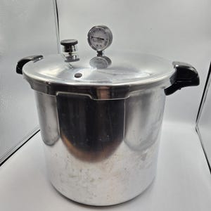 May include: A large, silver pressure cooker with a black handle and a pressure gauge on top. The cooker has a shiny, metallic finish and a round shape. The gauge has a white face with black markings. The cooker is designed for cooking food under pressure.