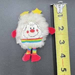 May include: A vintage plush toy with a white, fluffy body, a smiling face with a red nose, and a rainbow stripe. It has red hands and feet, and yellow star accents. The toy is approximately 3 inches tall.