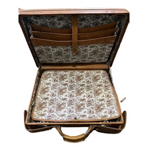 May include: Open brown leather briefcase with a handle and brass hardware. The interior features multiple pockets and a paisley patterned lining in shades of cream, brown, and red. The briefcase is designed to hold documents and other items.