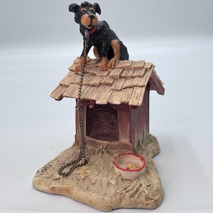 May include: A decorative figurine of a dog sitting on a dog house. The dog is black and brown with a red collar. The dog house is brown and red with a chain attached. A small bowl is at the base of the dog house.