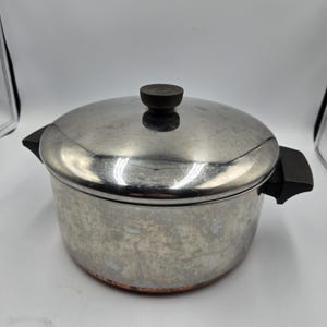 May include: A stainless steel cooking pot with a lid and two black handles. The pot has a copper-colored bottom and a black knob on the lid. The pot is designed for cooking and food preparation.