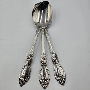 May include: Three silver spoons with ornate floral and scrollwork detailing on the handles. The spoons are arranged on a white surface, with the spoon bowls overlapping. The spoons are likely part of a vintage silverware set.