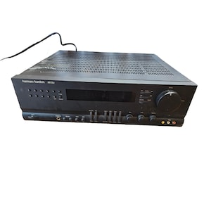May include: A black Harman Kardon AR 25 II stereo receiver. The front panel features a display, numerous buttons, knobs, and input jacks. The top has a perforated design for ventilation, and a power cord extends from the back.