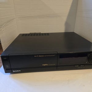 May include: Black Sony Hi-Fi Stereo VCR. The VCR has a power button, a disc symbol, and a control dial. The front of the VCR has the words "Hi-Fi Stereo" and "vcrplus+". The VCR is on a white surface.