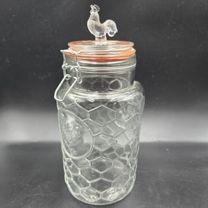 May include: A clear glass jar with a rooster-shaped lid. The jar has a honeycomb pattern and a hinged metal clasp. The lid has an orange rubber seal. The rooster is clear and sits atop the lid.
