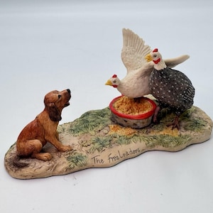 May include: A ceramic figurine depicting a brown dog looking up at two chickens near a bowl of feed. One chicken is white, the other is speckled. The base is inscribed with "The freeloaders."