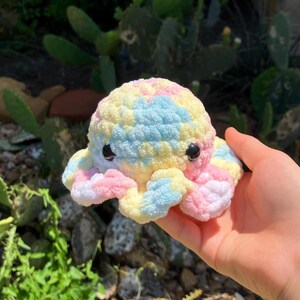 May include: A colorful, crocheted octopus toy in pastel shades of yellow, blue, and pink. The plush toy has black button eyes and is held in a hand. The background features green plants.