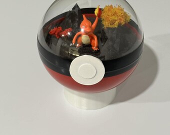 May include: A red, black, and white terrarium in the shape of a Poké Ball. Inside, a small orange figure of a Charmander stands before a miniature landscape with black mountains and orange and yellow foliage. The terrarium sits on a white base.