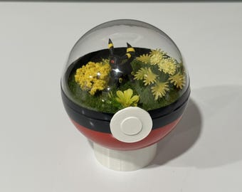 May include: A decorative terrarium in a Poké Ball design. The clear dome reveals a miniature scene with a black and yellow figure, yellow flowers, and green grass. The ball is red, black, and white, and sits on a white base.