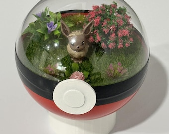 May include: A terrarium inside a red, black, and white sphere, featuring a small brown creature figurine, green grass, and colorful miniature plants. The sphere is mounted on a white base.