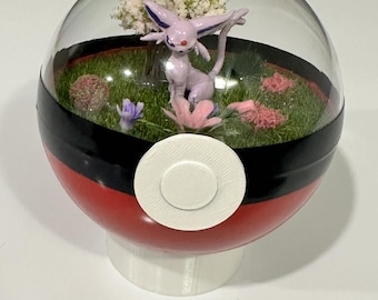 May include: A decorative terrarium inside a red, black, and white sphere. The terrarium features a purple Espeon figurine, flowers, and a small tree on a bed of green grass. The sphere is mounted on a white base.