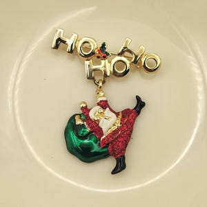 May include: Gold-coloured Christmas brooch with the words "HO HO HO" above a Santa Claus figure. Santa is in a red glittery suit, black boots, and a white beard, holding a green sack.