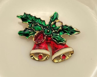 May include: A Christmas-themed brooch featuring two red bells with white trim and gold accents. The bells are adorned with a green holly leaf design. Each bell has a small red jewel at the center.
