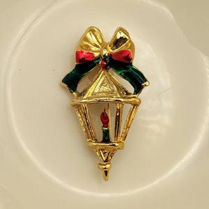 May include: A gold-coloured Christmas lantern brooch featuring a red candle and a bow with red and green details. The lantern has a triangular top and a decorative base. The brooch is set against a white background.