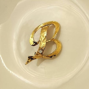 May include: A gold-colored metal brooch in the shape of the letter "B". The brooch has a textured surface and a small, clear gemstone. The brooch is set against a white background.