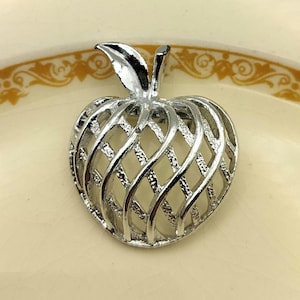 May include: Silver-coloured apple-shaped brooch with a lattice design. The brooch has a detailed leaf and stem, and the body of the apple is crafted with an open, woven pattern. This vintage-style brooch is a unique accessory.