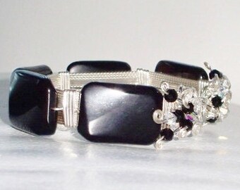 Puede incluir: Un brazalete negro y plateado con cuentas cuadradas negras y alambre plateado envuelto alrededor. El brazalete está adornado con cuentas de cristal transparentes y negras.