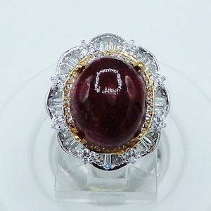 May include: A silver ring with a large, oval-shaped, dark red gemstone surrounded by a gold setting and smaller clear gemstones.