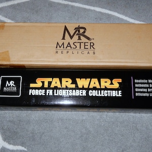 Peut inclure: Une boîte en carton marron avec le logo "MR MASTER REPLICAS" au-dessus d'une boîte noire avec le texte "STAR WARS FORCE FX LIGHTSABER COLLECTIBLE". Comprend une poignée en métal et une lame violette brillante.
