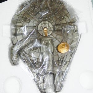 Puede incluir: Un modelo detallado, de color plateado, de la nave espacial Millennium Falcon, empaquetado en plástico transparente. El modelo presenta detalles intrincados y una pieza dorada. La nave está en una caja blanca.