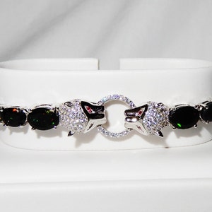 Puede incluir: Una pulsera de tono plateado con una serie de piedras negras ovaladas con motas iridiscentes. El diseño central consta de dos cabezas de pantera plateadas incrustadas con piedras claras, enfrentadas y sosteniendo un pequeño anillo.