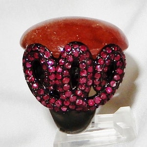 May include: A ring with a large, red gemstone set in a black metal band. The band is decorated with a swirling design of small, pink gemstones.