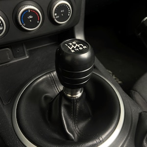May include: A black gear stick knob with white gear pattern markings, installed in a car. The shift knob is cylindrical with horizontal grooves. The surrounding interior is black leather and plastic.